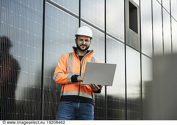 Smiling engineer holding laptop standing near solar panels