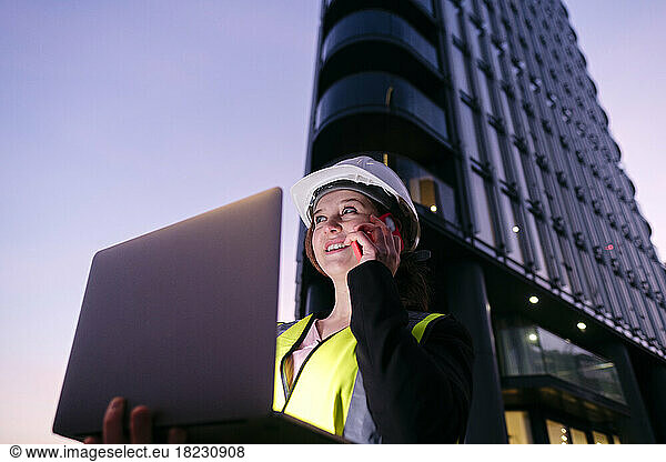 Smiling engineer holding laptop talking on mobile phone in front of building