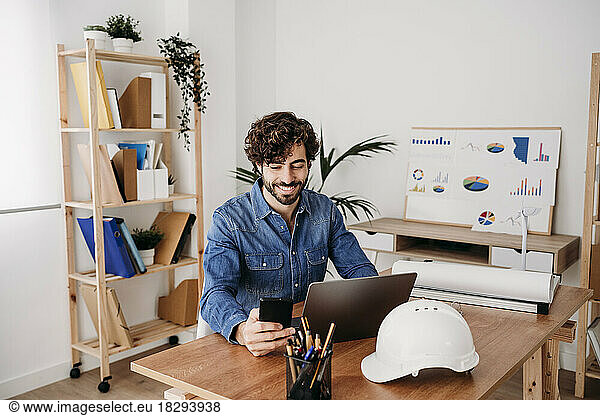 Smiling engineer using smart phone with laptop and hardhat on desk in office