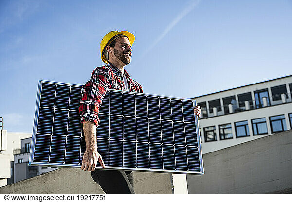 Smiling engineer wearing hardhat carrying solar panel