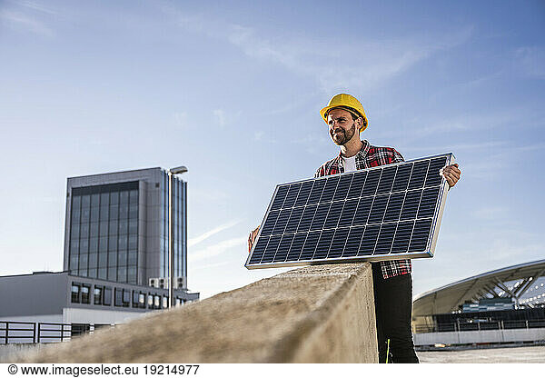 Smiling engineer wearing hardhat holding solar panel at terrace on sunny day