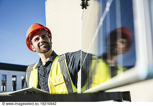 Smiling engineer wearing protective workwear holding digital tablet examining solar panel at terrace on sunny day