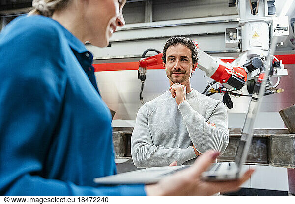 Smiling engineer with hand on chin looking at colleague in robot factory