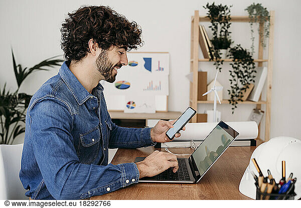 Smiling engineer working on laptop and smart phone sitting at desk in office