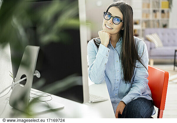Smiling entrepreneur wearing eyeglasses sitting with hand on chin by desk at office