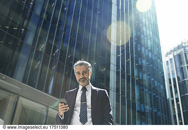 Smiling entrepreneur with mobile phone outside office building in city during sunny day