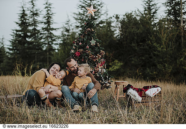 Smiling family sitting by Christmas tree on grassy land at countryside during sunset