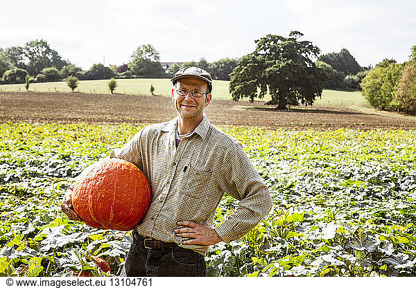 Smiling farmer standing in a field  holding large red pumpkin.