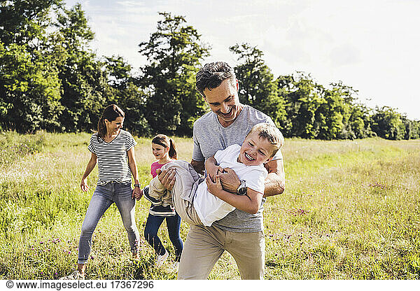 Smiling father carrying son while walking with family on meadow