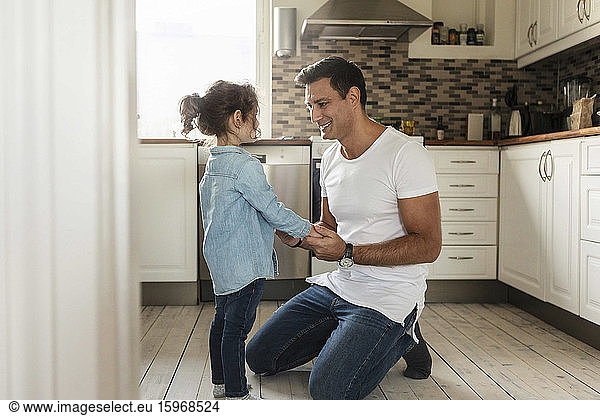 Smiling father holding hands with daughter while kneeling on kitchen floor