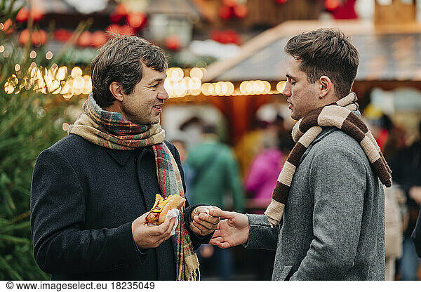 Smiling father holding hot dog and talking with son standing at Christmas market