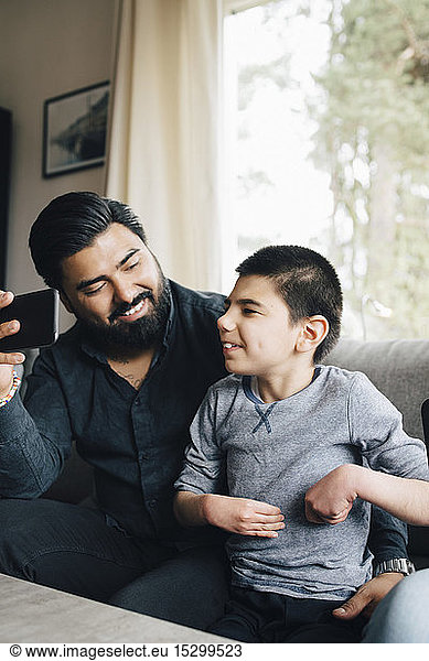Smiling father holding mobile phone looking at autistic son while sitting on sofa in living room