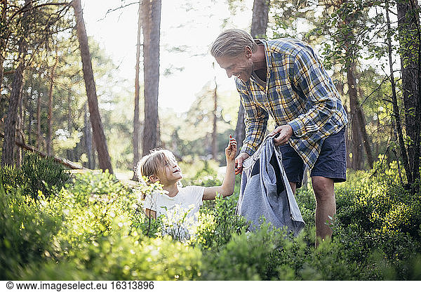 Smiling father looking at daughter collecting garbage in forest