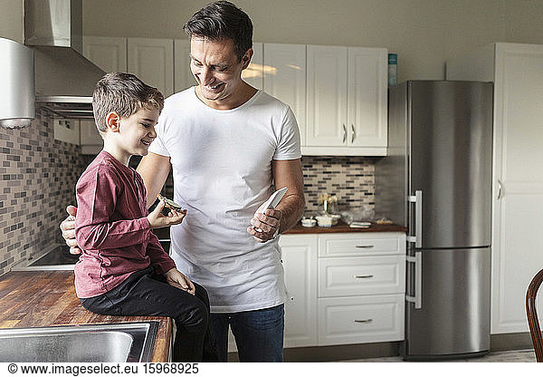 Smiling father showing smart phone to son with food while standing by kitchen counter