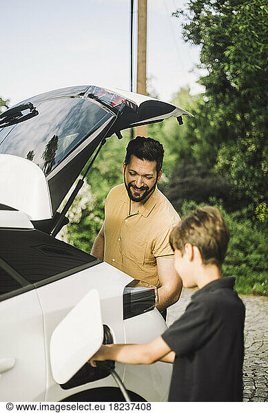 Smiling father standing by son charging electric car