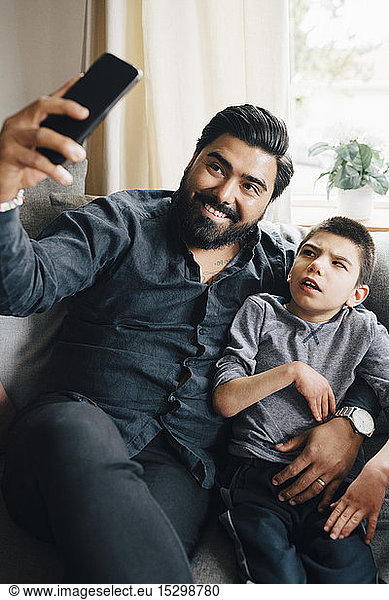 Smiling father taking selfie with autistic son while sitting on sofa in living room