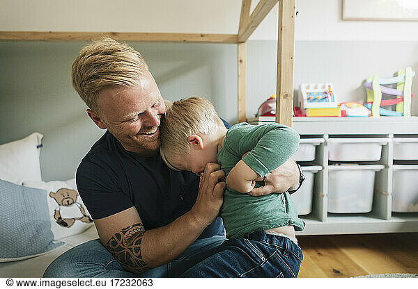 Smiling father tickling son while sitting in bedroom