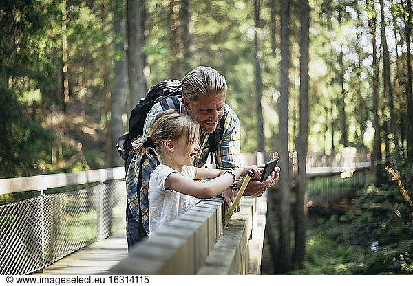 Smiling father with backpack and daughter looking at smart phone in forest