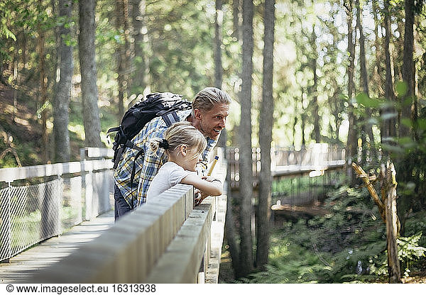 Smiling father with backpack talking to daughter on footbridge in forest