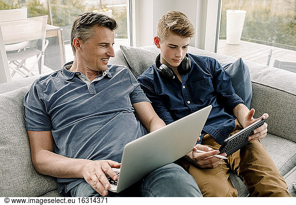 Smiling father with laptop looking at son using digital tablet while sitting on sofa at home