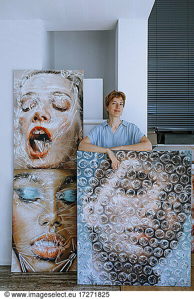 Smiling female artist standing with paintings in art studio
