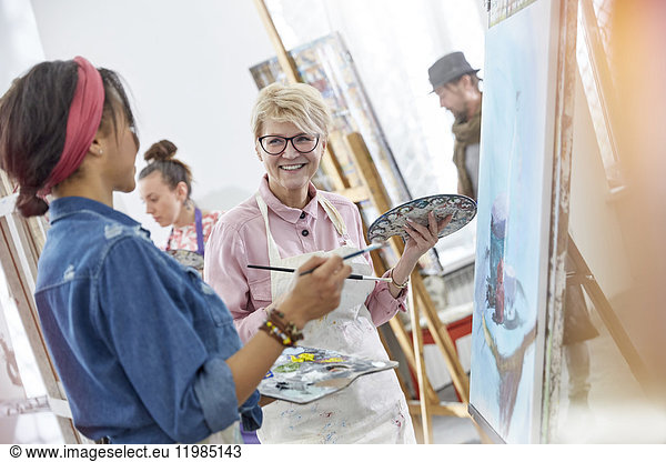 Smiling female artists with paintbrushes and palettes painting in art class studio