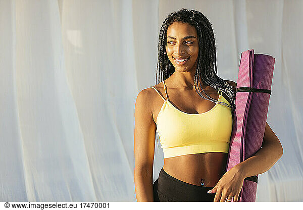 Smiling female athlete looking away while holding exercise mat