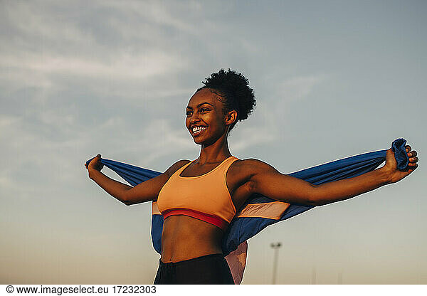 Smiling female athlete with arms outstretched against sky