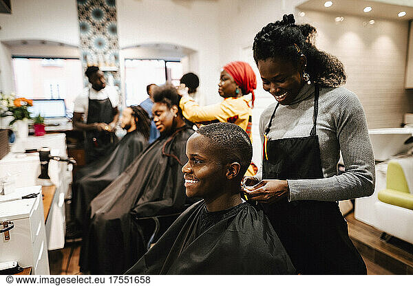 Smiling female barber making locs of male customer in salon