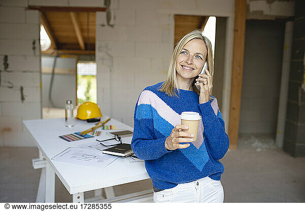 Smiling female building contractor with disposable coffee cup talking on mobile phone while leaning on table at site