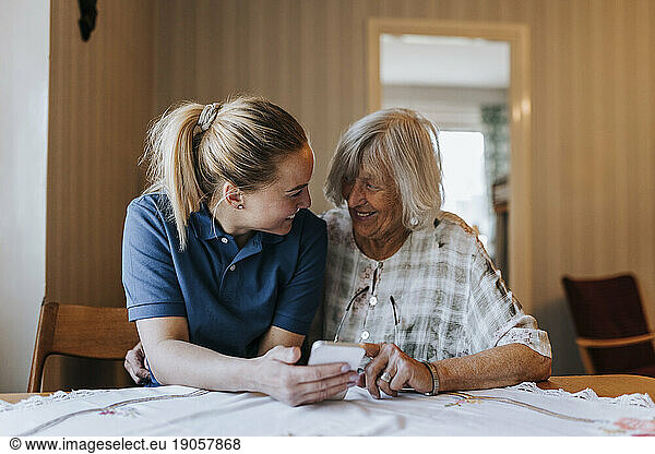 Smiling female care assistant sharing smart phone with senior woman while sitting at dining table in home