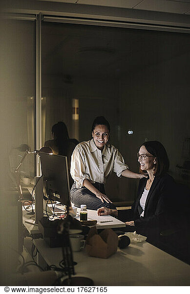 Smiling female colleagues discussing on computer while working last minute at work place