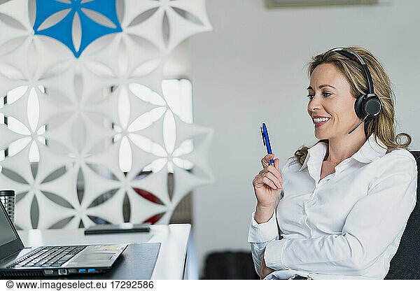 Smiling female customer service representative with pen talking through headphones at desk in office