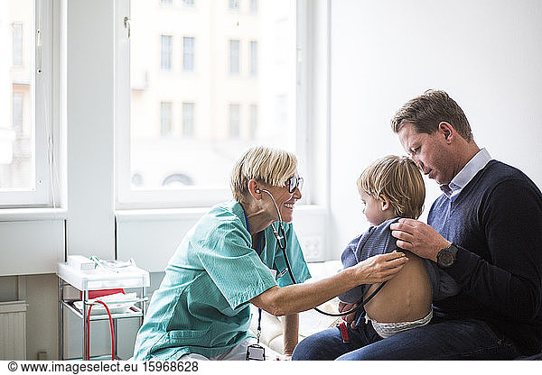 Smiling female doctor examining boy's back with stethoscope while sitting by father in hospital
