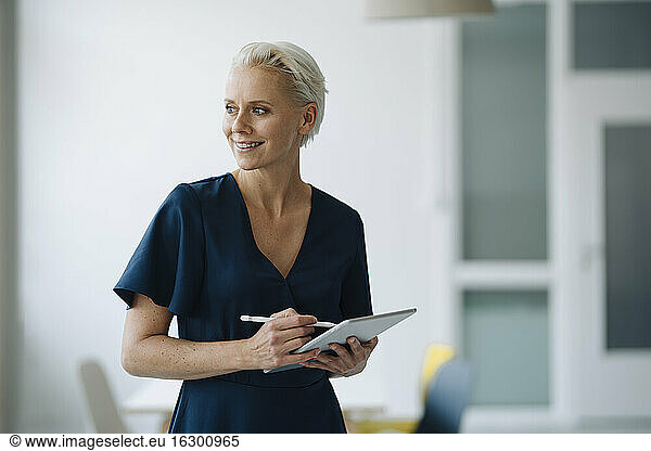 Smiling female entrepreneur holding digital tablet contemplating while standing in office