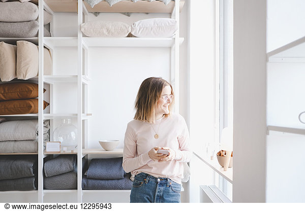 Smiling female entrepreneur holding mobile phone while looking through window in upholstery store