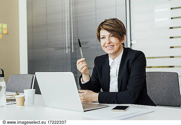 Smiling female entrepreneur holding pen in front of laptop at desk in office