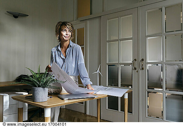 Smiling female entrepreneur looking away while holding blueprint sitting on desk in office