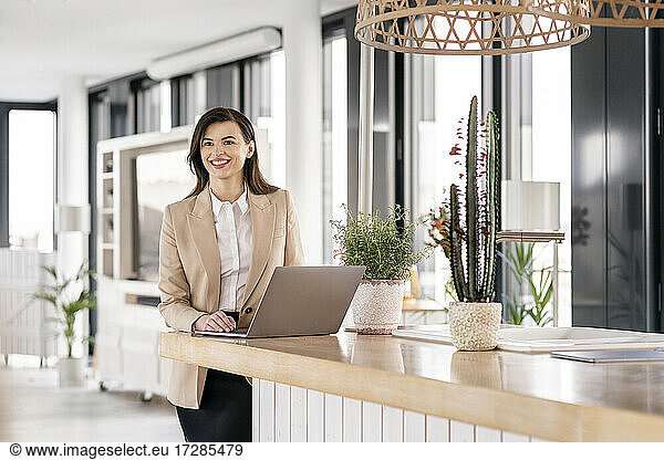 Smiling female entrepreneur looking away while standing in front of laptop at office cafeteria