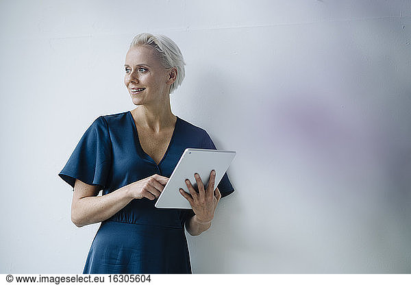 Smiling female entrepreneur looking away while working over digital tablet against wall in office