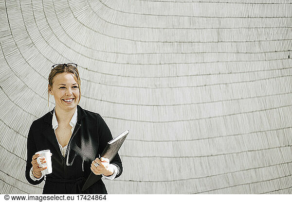 Smiling female entrepreneur standing with coffee mug and file at convention center looking away