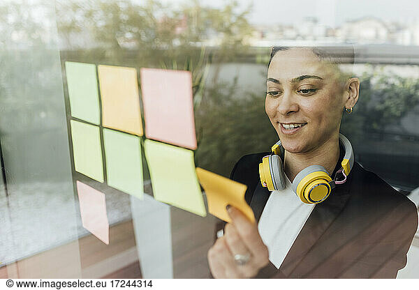 Smiling female entrepreneur sticking adhesive notes on glass in office