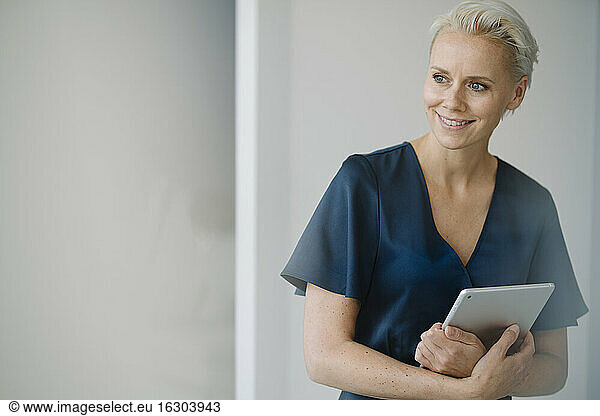 Smiling female entrepreneur with digital tablet looking away while standing against wall in office