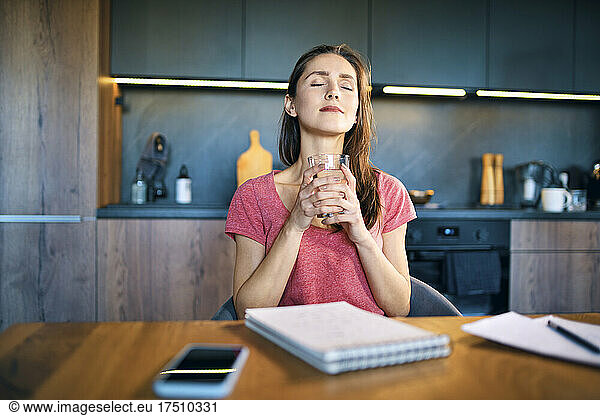 Smiling female entrepreneur with eyes closed holding drinking glass at desk in home office