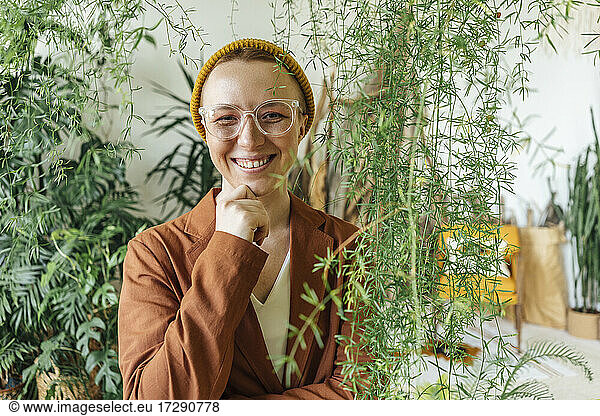 Smiling female entrepreneur with hand on chin standing by plants at work place