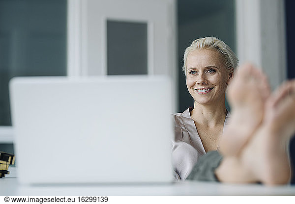 Smiling female entrepreneur with laptop on desk contemplating while relaxing in office