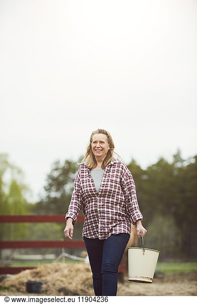 Smiling female farmer holding bucket while walking on field against sky