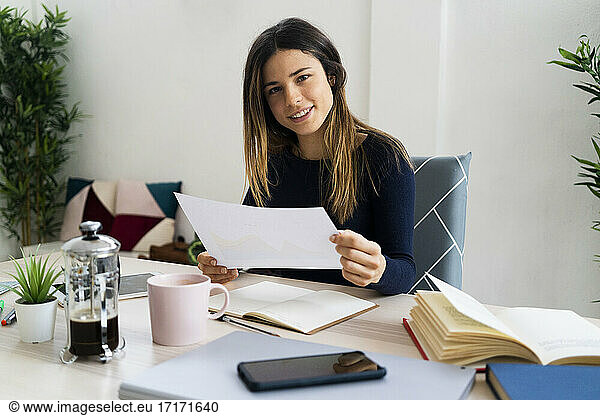 Smiling female freelance worker holding paper while sitting on chair at home
