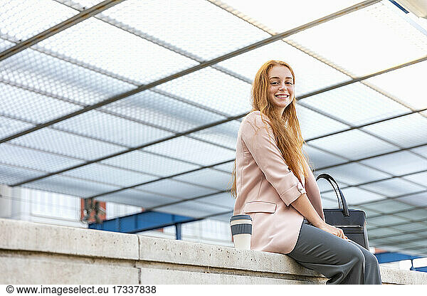 Smiling female freelancer sitting on retaining wall under shade at railway station