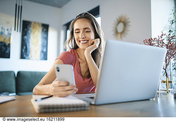 Smiling female freelancer using smart phone by laptop on desk in home office